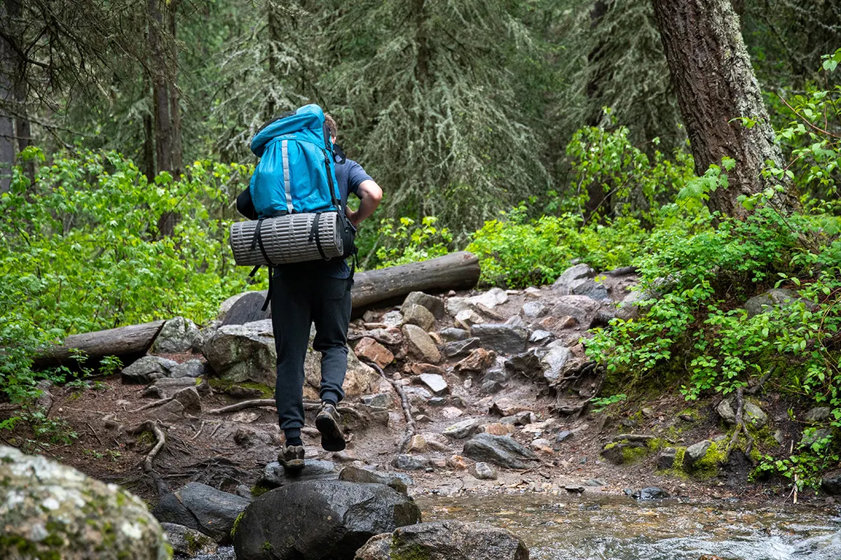 Hiker with a blue backpack and sleeping mat crossing a rocky forest stream trail