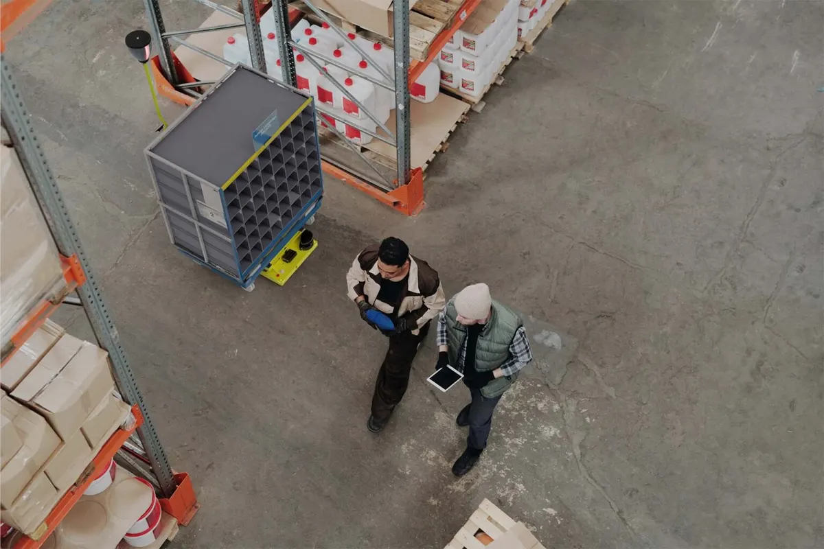 Overhead view of two warehouse workers walking past an IW.HUB carrying a shelving trolley, fitted with the Reflection Flag